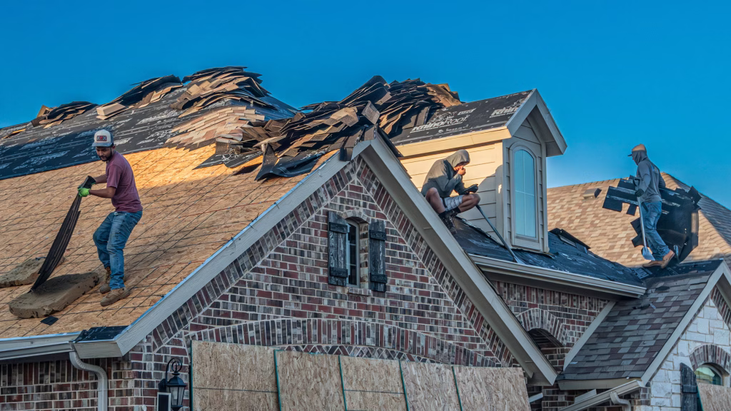 Roofing crew members removing old asphalt shingles from a steep residential roof on a brick home under a clear blue sky.