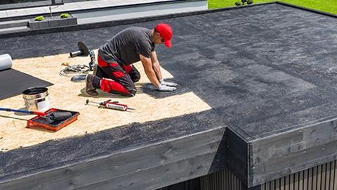 A construction worker applies EPDM roofing material on a flat roof of a modern home in a sunny outdoor environment. Landscaping is visible in the background.