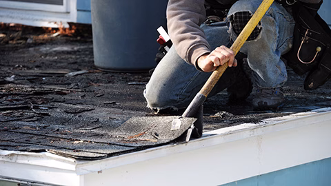 Roof construction worker removing old damaged shingles as first step of repair process.