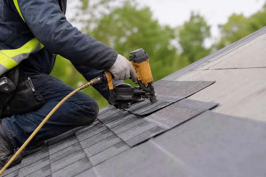 Roofing contractor securing asphalt shingles with a nail gun on a sloped residential roof during installation.