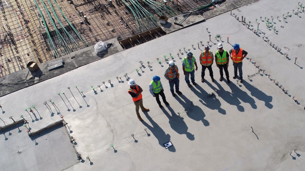 An aerial view of a professional roofing crew in safety vests and hard hats standing on a large concrete commercial rooftop during a construction project.
