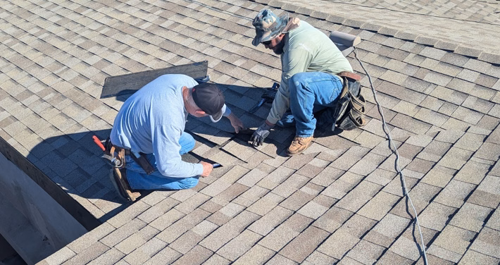 Two roofing contractors repairing damaged asphalt shingles on a sloped roof, replacing roofing material around a repair area.